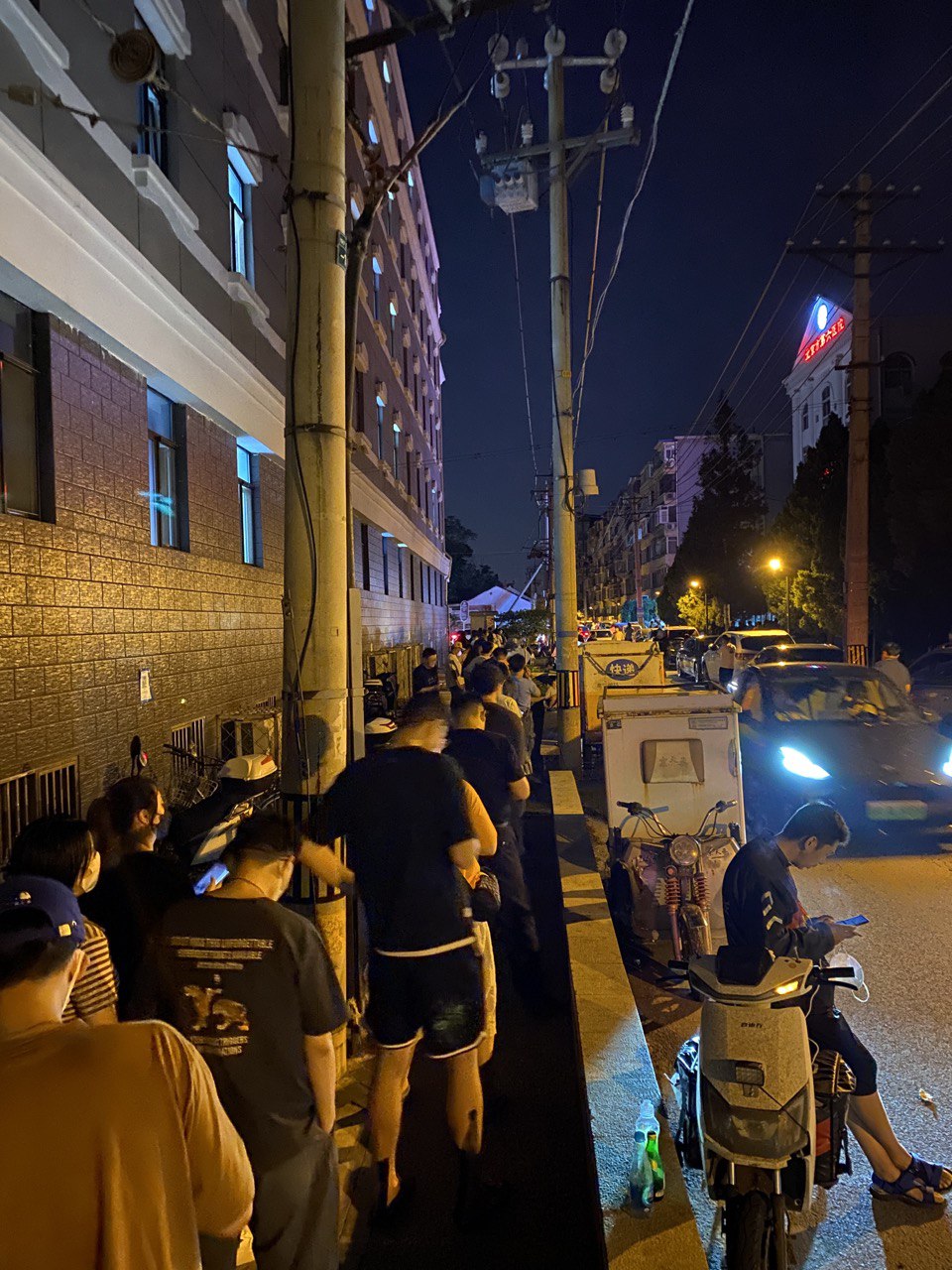 一张在夜晚拍摄的街景照片：较窄的小巷中有等候进行 COVID-19 病毒检测的队伍，以及多个行人与车辆，右侧的霓虹灯上的文字是“北京市第六医院”