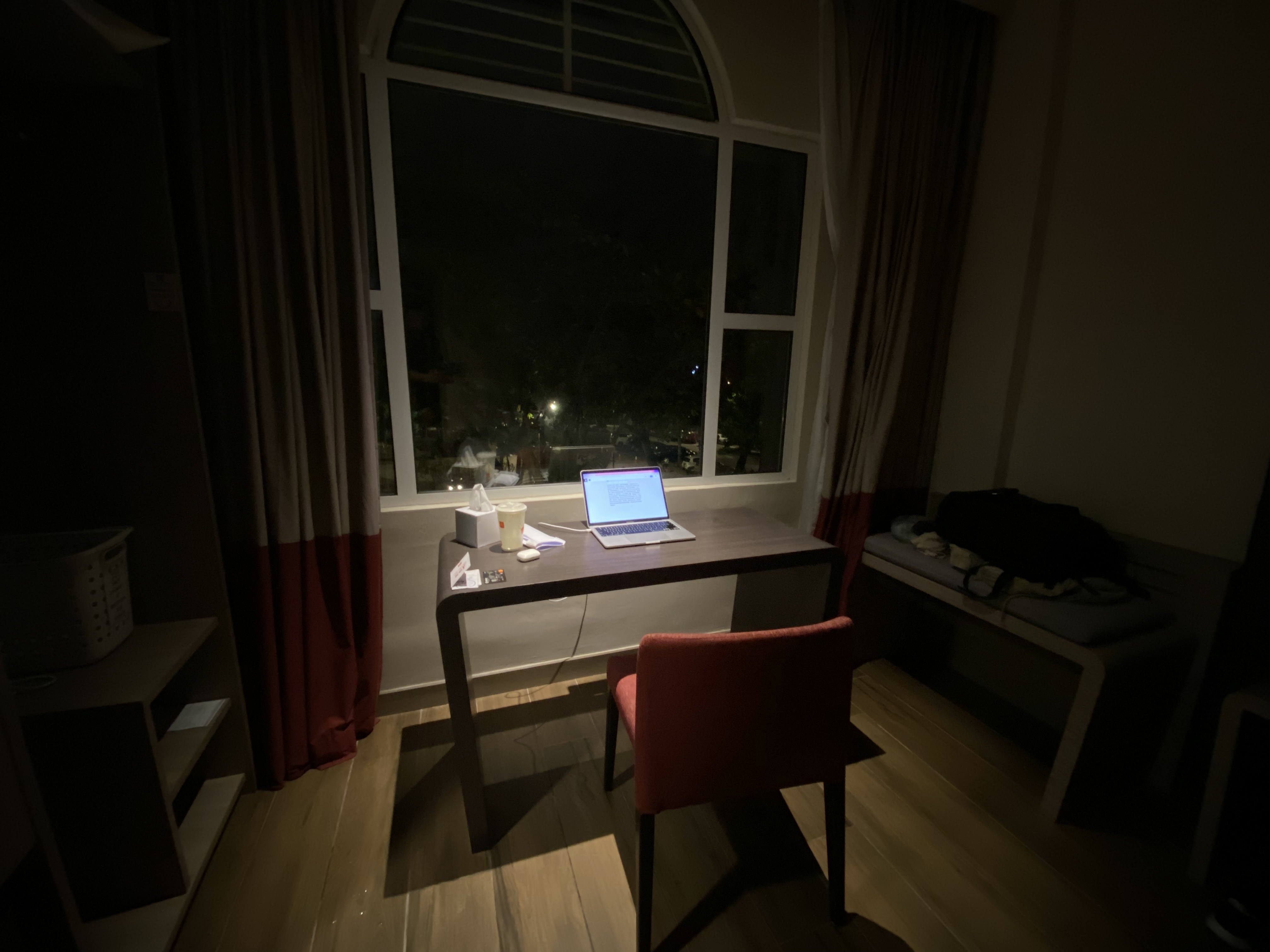 一张酒店房间中拍摄的图片，展示了一个只有面向窗户的书桌被投影灯光的场景。书桌上有餐巾纸、饮料、笔记本计算机和几张卡片